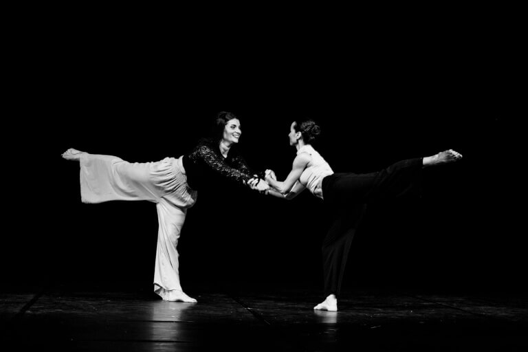 Deux danseuses en interaction, gestes synchronisés sur une scène sombre, lumière concentrée sur elles
