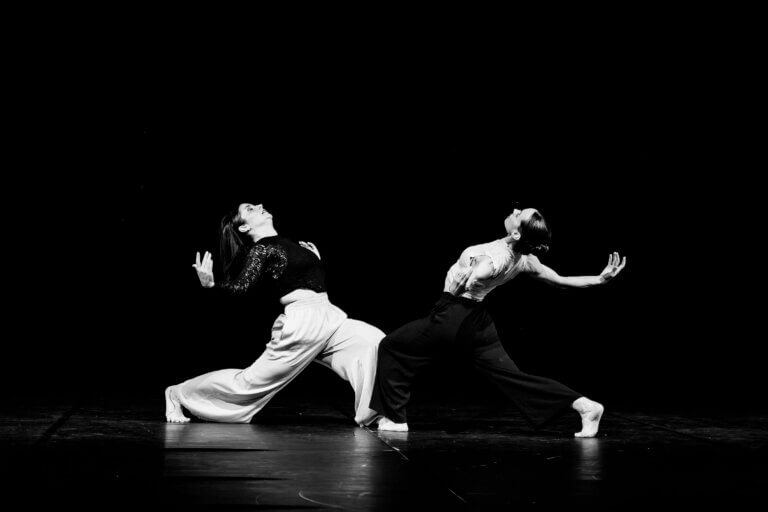 Deux danseuses en interaction, gestes synchronisés sur une scène sombre, lumière concentrée sur elles