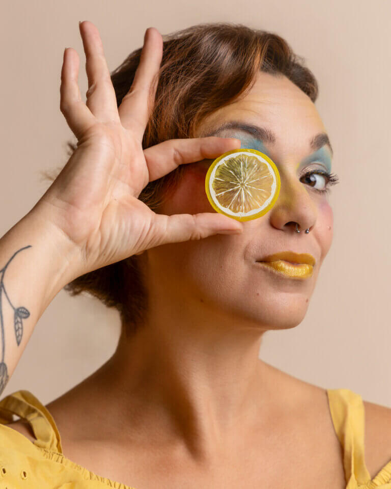 Portrait créatif d’une femme tenant une tranche de citron devant son œil avec un maquillage jaune et bleu.