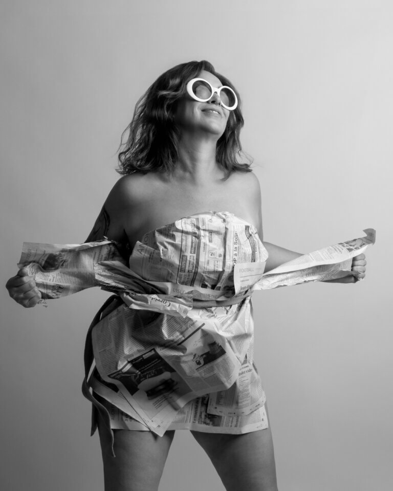 Portrait créatif en noir et blanc d’une femme portant une tenue en papier journal et des lunettes blanches.