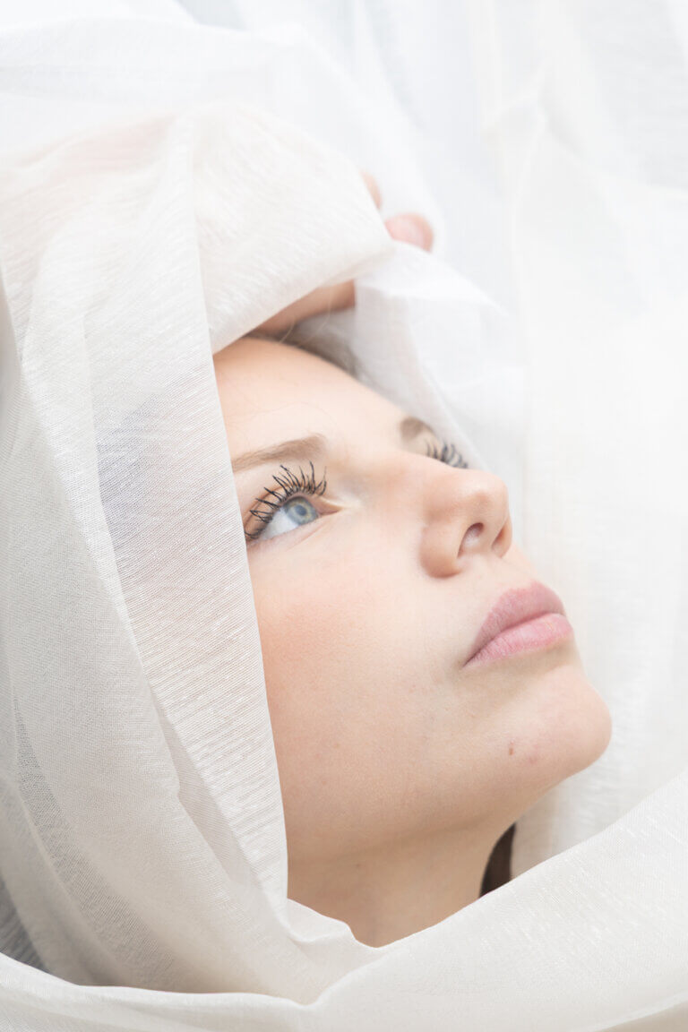 Portrait rapproché d’une femme dont le visage apparaît entre plusieurs voiles blancs transparents.