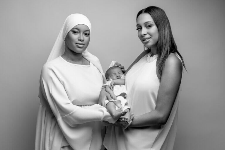 Deux soeurs tenant un nouveau-né endormi lors d’un portrait de famille en noir et blanc.